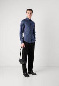 Cămașă cu dungi navy, cu butoni și mâneci lungi, purtată cu pantaloni negri și pantofi negri din piele. Ține o geantă mică de umăr neagră.
