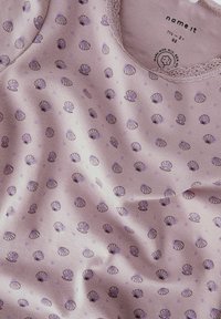 Jasnofioletowa koszula z krótkim rękawem, wykonana z miękkiego materiału, zdobiona wzorem muszelek w ciemniejszym fiolecie. Ozdobne koronkowe wykończenie wokół dekoltu.