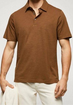 Mand iført en brun kortærmet polo t-shirt med to knapper, kombineret med lyse beige bukser, holder en hvid beklædningsgenstand i venstre hånd.