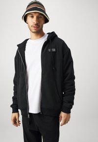 Melna zip-up džempera ar ribinātu apkakli un taisnstūra logo uzlīmi, kombinēts ar balto T-kreklu. Modelis valkā svītrainu bļodiņu cepuri.