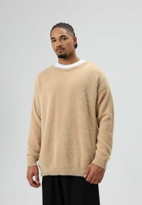 Beige gebreide trui van zacht materiaal met een losse pasvorm, een brede halslijn en lange mouwen, gecombineerd met een zwarte broek.