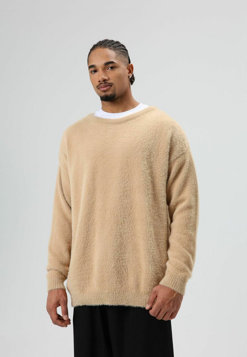 Beige gebreide trui van zacht materiaal met een losse pasvorm, een brede halslijn en lange mouwen, gecombineerd met een zwarte broek.