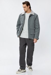 Veste isolante grise avec un col en fausse fourrure gris clair, poches avant, associée à un pantalon cargo noir et des baskets beiges.