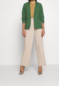 Green blazer with notched lapels, paired with a mustard-colored top and beige wide-leg trousers. Accented with snake-print high heels.