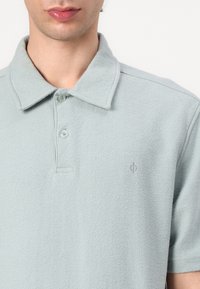 Gros plan d'une personne portant un polo bleu clair texturé avec un col et deux boutons, montrant la partie supérieure de la poitrine et du cou.