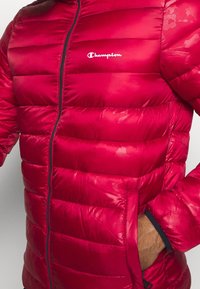 Rød puffjakke med lynlås foran, vandret syninger og et lille Champion-logo på brystet. Har sidelommer og en glansfuld overflade.