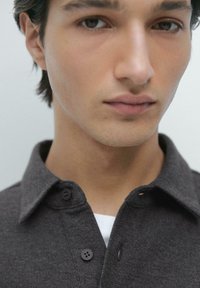 Grijs overhemd met knopen en een gestructureerde stof, voorzien van een kraag en twee knopen aan de voorkant. Witte ondershirt gedeeltelijk zichtbaar eronder.