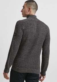Strikket mørkegrå sweater med høj krave, ribbede manchetter og kant. Indeholder et lille sort logo-patch på venstre ærme.