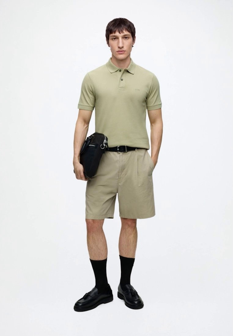 Jeune homme debout portant un polo vert clair, un short beige, une ceinture noire, des chaussettes noires, des chaussures noires, et tenant un sac noir d'une main.