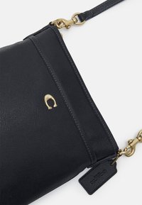 Černá kožená crossbody taška s texturovaným povrchem, zlatými kovovými doplňky a visačkou. Obsahuje zipové uzavření a dekorativní logo.