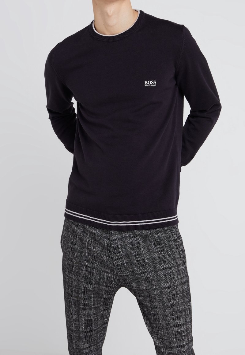 Mann trägt einen schwarzen Hugo Boss Pullover mit langen Ärmeln und weißem Logo sowie schwarze und weiße gemusterte Hosen und steht vor einem schlichten hellen Hintergrund.