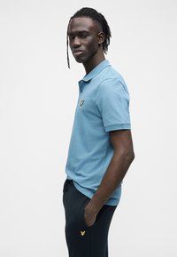 Lyseblå polotrøye med klassisk krage, korte ermer og en gullbrodert logo på brystet, kombinert med mørke marineblå bukser.