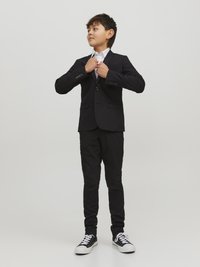 Svart skräddarsydd kostym med en knapp, vit skjorta under, slim-fit svarta byxor och svarta sneakers med vita sulor.