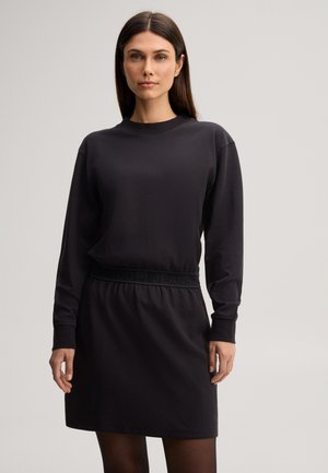 Donna con capelli lisci scuri che indossa un vestito nero a maniche lunghe, in piedi contro uno sfondo chiaro e uniforme.