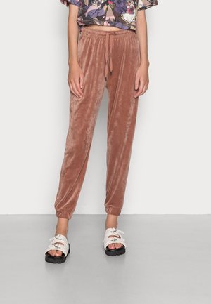 Woman wearing rust-colored velour joggers with drawstring waist and white open-toe sandals standing on gray floor.