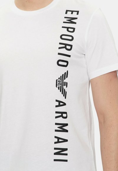 T-shirt bianca in cotone con testo verticale nero che recita "EMPORIO ARMANI" e un piccolo logo ad aquila. Maniche corte e scollatura a girocollo.