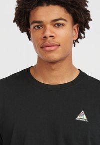 Black cotton crew neck t-shirt featuring a small triangular logo patch with colorful design on the left chest area. Smooth texture.