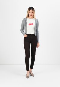 Svijetlosivi zip-up kardigan preko bijele majice s crvenim grafičkim motivom trešnje, u kombinaciji s uskim crnim trapericama i sivim balerinkama.