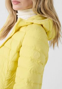 Giacca a vento gialla con design trapuntato, dotata di cappuccio e logo in rilievo sulla manica. Materiale morbido e leggero con cuciture ben visibili.