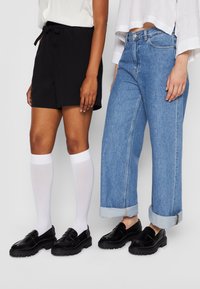 Shorts neri in cotone con vita annodata e calze bianche fino al ginocchio, abbinati a jeans in denim blu chiaro con orli arrotolati e mocassini neri.