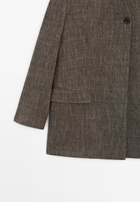 Blazer brun texturé à manches longues, avec un bouton noir et une poche à rabat horizontale sur fond blanc.