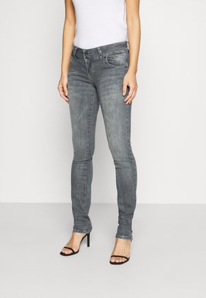 Vrouw draagt vervaagde grijze slim-fit jeans en zwarte open teen hakken en staat tegen een effen lichte achtergrond.