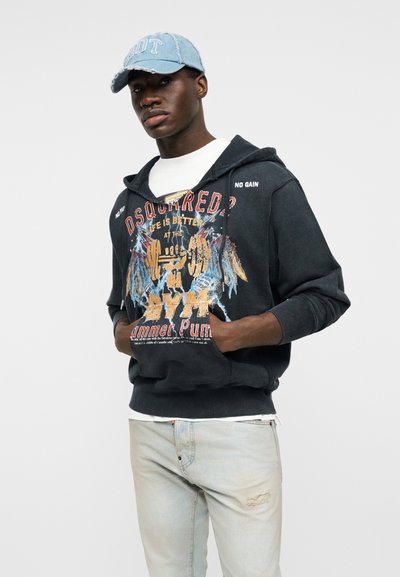 Jeune homme portant une casquette bleue, un sweat à capuche noir avec un motif sur le thème de la salle de sport, un t-shirt blanc et un jean clair déchiré, debout les mains dans les poches.