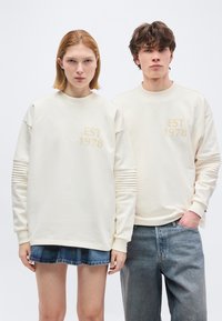 Νεαρή γυναίκα και άνδρας που φοράνε ταιριαστά κρεμ φούτερ με το κείμενο "EST 1978", η γυναίκα με φούστα από τζιν, ο άνδρας με μπλε τζιν, στέκονται δίπλα δίπλα.