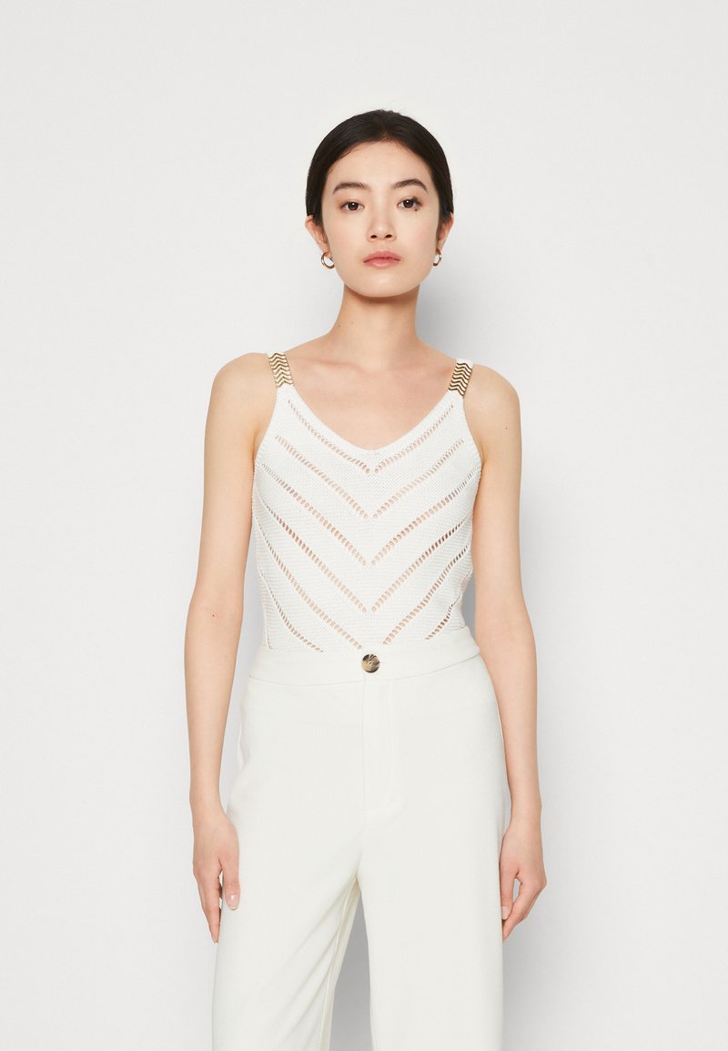 Witte tanktop met een zigzag haakpatroon en gouden schouderbanden, gecombineerd met witte high-waisted broek met knoopdetails.