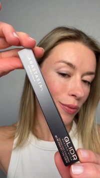 Woman holding Anastasia Beverly Hills Glider Shadow Stick makeup product close to her face against a plain background.