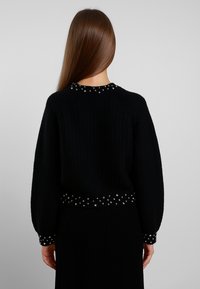 Maglione nero lavorato a maglia con texture a coste, presenta borchie argento lungo il collo e i polsini, e una vita aderente.