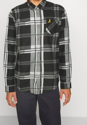 Czarna, biała i szara koszula w kratę z guzikami i kieszenią na piersi z małym żółtym logo ptaka, noszona na wierzchu ciemnych spodni.