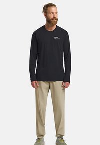 Maglietta nera a maniche lunghe con logo sul petto, abbinata a pantaloni beige chiaro e scarpe da ginnastica grigio-verdi. Il tessuto appare liscio e leggero.