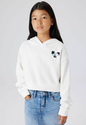 Sweat à capuche court blanc avec une poche avant, affichant un petit motif de cœur noir et pastel. Associé à un short en denim taille haute.
