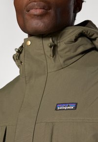 Oliven grøn Patagonia jakke med lynlås og trykknaplukning, justerbare snore, og et logo patch på brystet. Glat stof texture.