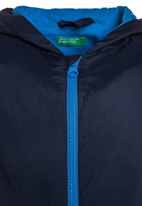 Chaqueta azul marino con cremallera y capucha en azul brillante, con una etiqueta verde "United Colors of Benetton" dentro del collar.