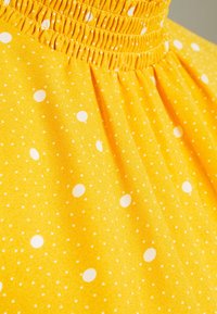 Gult tyg med vita prickar; har en skrynklig halsringning och en slät yta i en mjuk, strömmande design.