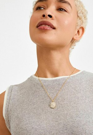 Goudkleurige halsketting met een gestructureerd hangertje, gedragen over een grijze mouwloze top. Het hangertje heeft een fonkelend, gedetailleerd ontwerp.