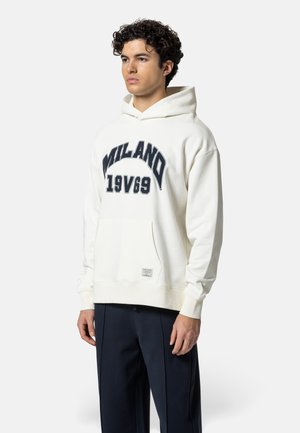 Junger Mann mit lockigem Haar, der einen weißen Hoodie mit dem Text "MILANO 19V69" und marineblaue Hose trägt, vor einem schlichten Hintergrund stehend.
