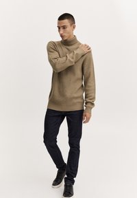 Beige ribbade turtleneck-sweater med långa ärmar, kombinerat med mörkblå slim-jeans och svarta sneakers, uppvisat mot en ljus bakgrund.
