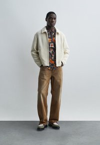 Νέος άνδρας που στέκεται με τα χέρια στις τσέπες, φορώντας κρεμ πλεκτό μπουφάν, πορτοκαλί και καφέ πουκαμίσα με σχέδια, καφέ φαρδιά παντελόνια και δίχρωμα παπούτσια.