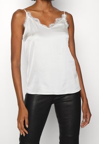 Camiseta de satén blanca con tirantes finos, que presenta un delicado encaje en el escote. Textura suave y diseño ajustado. Se lleva con pantalones de cuero negros.