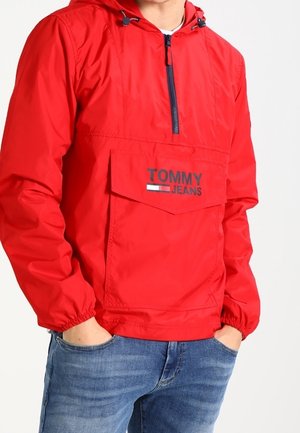 Mann trägt eine rote Tommy Jeans Kapuzen-Windjacke mit Fronttasche und blaue Jeans, Hände in den Taschen, vor weißem Hintergrund.