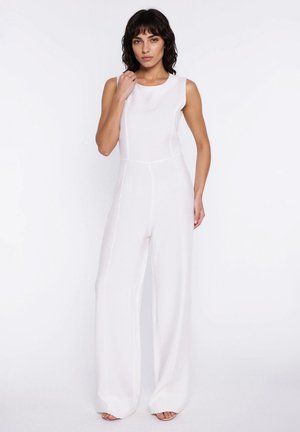 Kobieta o ciemnych włosach ubrana w bezrękawnik i szerokie, białe kombinezon, stoi na białym tle.