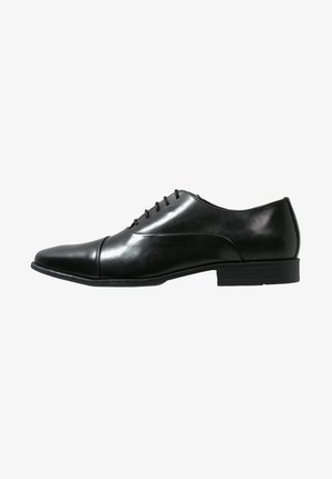 Chaussure de ville en cuir noir avec un bout pointu, six œillets pour les lacets, texture lisse et un talon bas. Design simple et classique.