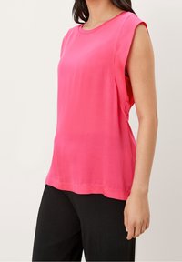 Růžový bezrukávový top s kulatým výstřihem, hladkým povrchem a volným střihem. Zadní lem je mírně delší, doplněný černými kalhotami.