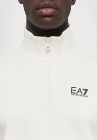Hvit zip-up hettegenser med høy krage, med en svart EA7 Emporio Armani-logo på venstre side og en teksturert stofffinish.