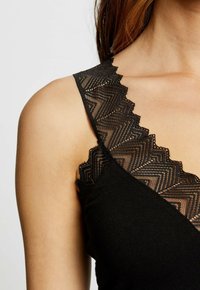Top nero senza maniche con un bordo in pizzo a scalloped lungo il colletto. Il pizzo presenta un motivo a zigzag, che completa il tessuto liscio.