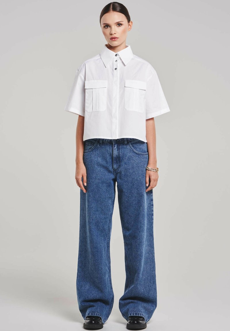 Cropped white button-up shirt with two chest pockets, paired with high-waisted, wide-leg blue denim jeans, finished with black footwear.