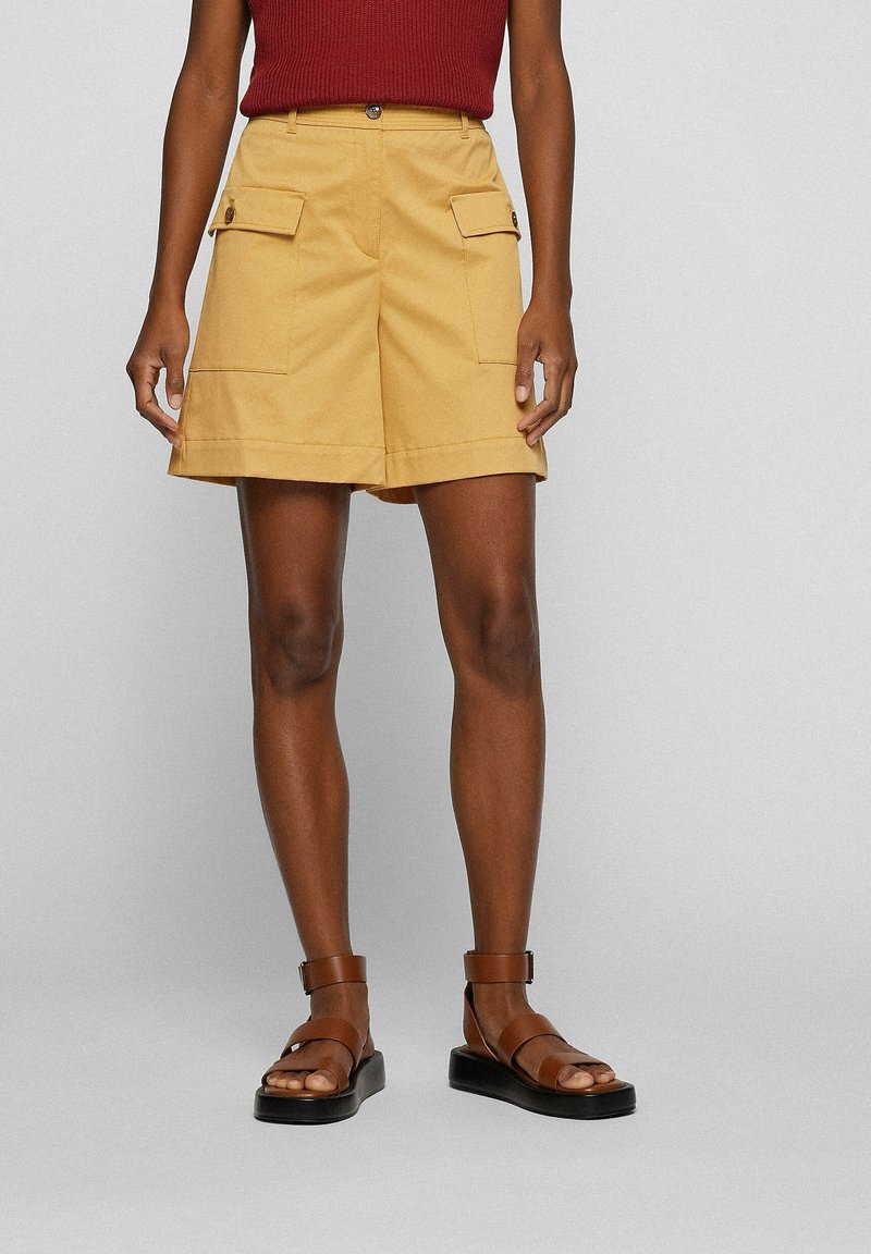Personne portant un short jaune moutarde longueur genou avec des poches à rabat à l'avant, un top rouge sans manches et des sandales marron à bride cheville.